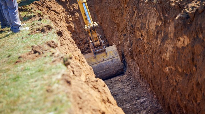 Yellow excavator digging a precise trench in a grassy field for utility or drainage installation.