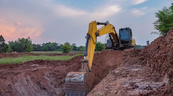 A yellow excavator digging a deep trench in red clay soil against a backdrop of a colorful sunset sky and distant trees.