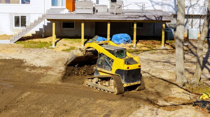 A yellow compact track loader moving soil in a backyard next to a house with a wooden deck, preparing the area for landscaping or drainage.