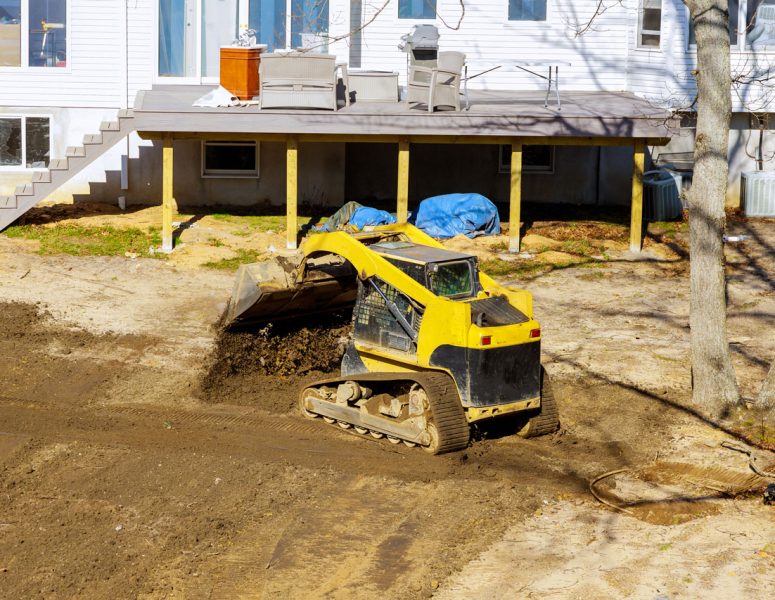 A yellow compact track loader moving soil in a backyard next to a house with a wooden deck, preparing the area for landscaping or drainage.