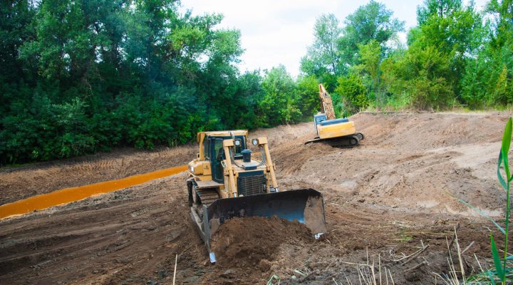 A bulldozer grading a slope next to a muddy drainage ditch, with an excavator working in the background amidst dense green trees.