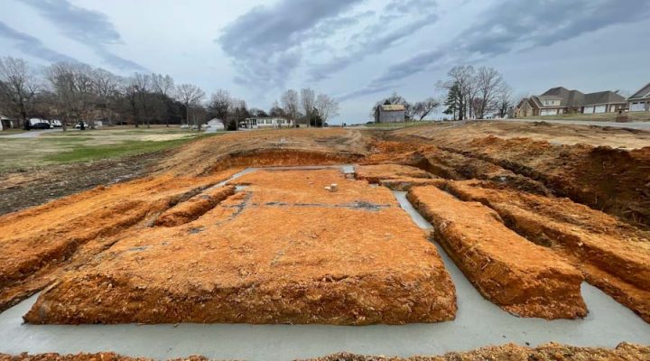 A detailed view of a home’s concrete foundation trenches filled with fresh concrete, surrounded by deep red clay soil under an overcast sky.