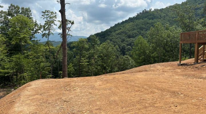 A wide, cleared hilltop lot with a wooden deck structure on the right, offering expansive views of lush green mountains under a partly cloudy sky.