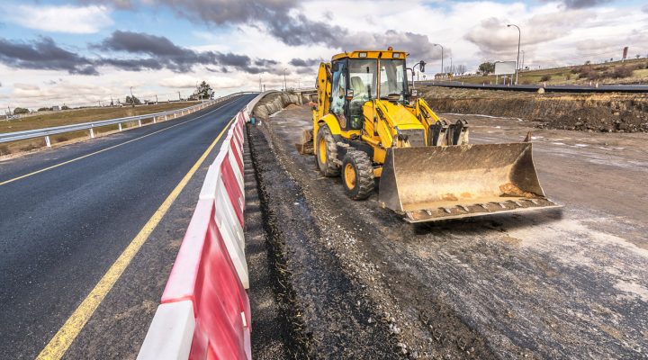 A yellow front-end loader working on a highway construction site next to a newly paved road, with red and white safety barriers in place.