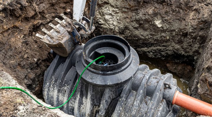 Close-up view of a black plastic septic tank component being installed in a muddy trench, with an excavator bucket positioned above.
